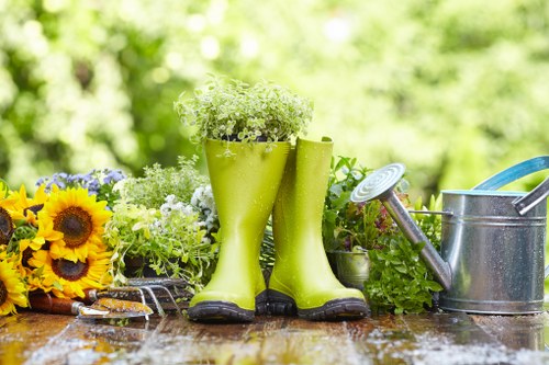 The image features a garden setting with a weathered wooden surface holding gardening tools and decorative elements. In the foreground, two bright yellow rubber boots are repurposed as planters filled with small, white flowering plants, adding a playful touch to the outdoor space. To the left, a bouquet of sunflower blooms and other mixed flowers with yellow, purple, and green foliage provides vibrant natural colour accents. On the right, there is a traditional metal watering can with a long spout and a handle, indicating active garden maintenance. The background reveals a lush, blurred garden with green trees and shrubbery, suggesting a well-maintained outdoor area typical of residential gardens in Hatch End. The lighting appears natural and bright, indicative of a clear day with sunlight filtering through the foliage, highlighting the textures of the plants and gardening equipment. This scene exemplifies a tidy, cultivated garden environment, possibly serviced by local gardening specialists like Hedge Trimming Hatch End, emphasising outdoor care and landscaping efforts for residential outdoor spaces in the area.